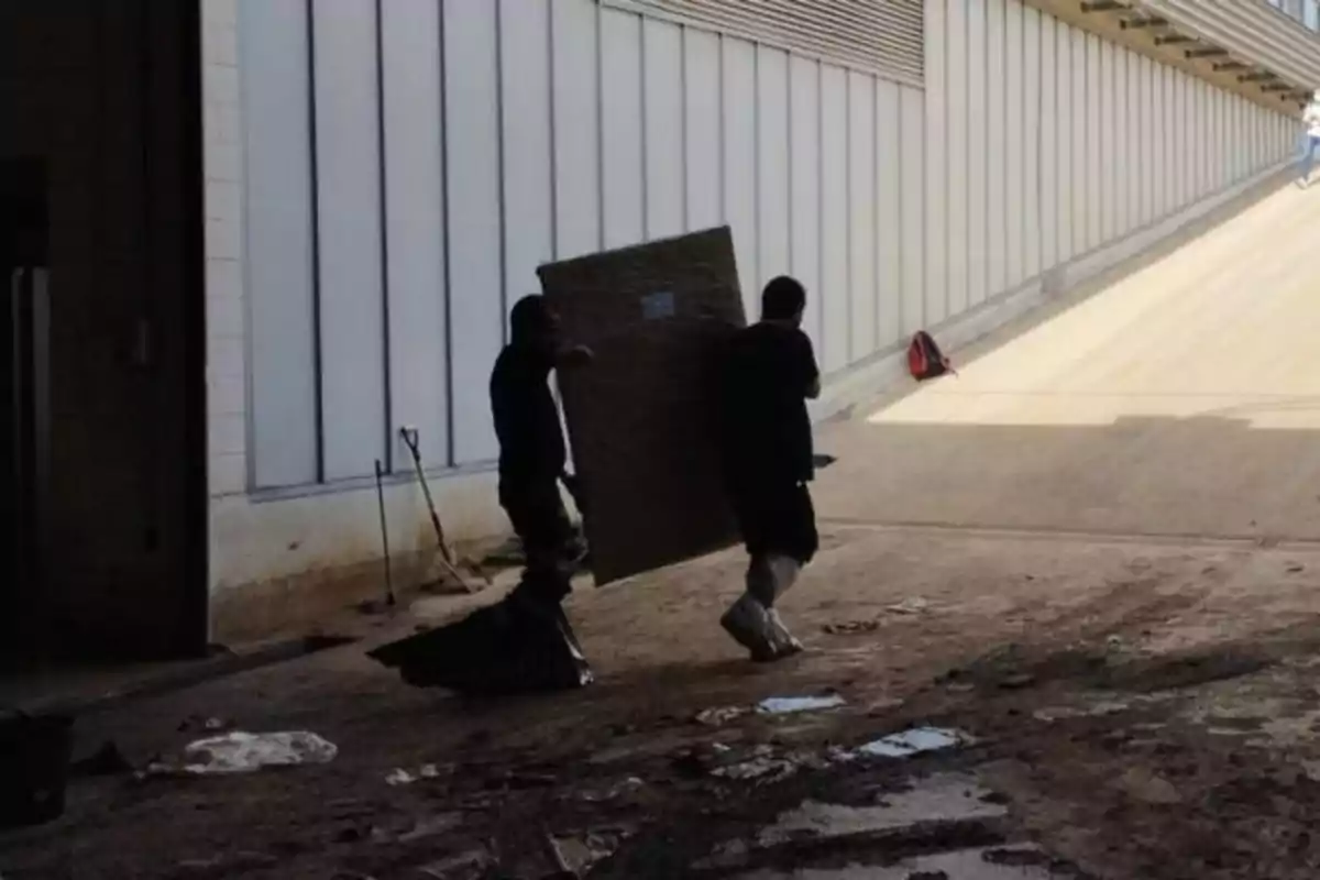 Dos personas cargan un objeto grande y rectangular mientras caminan por un área con suelo sucio y paredes de metal.