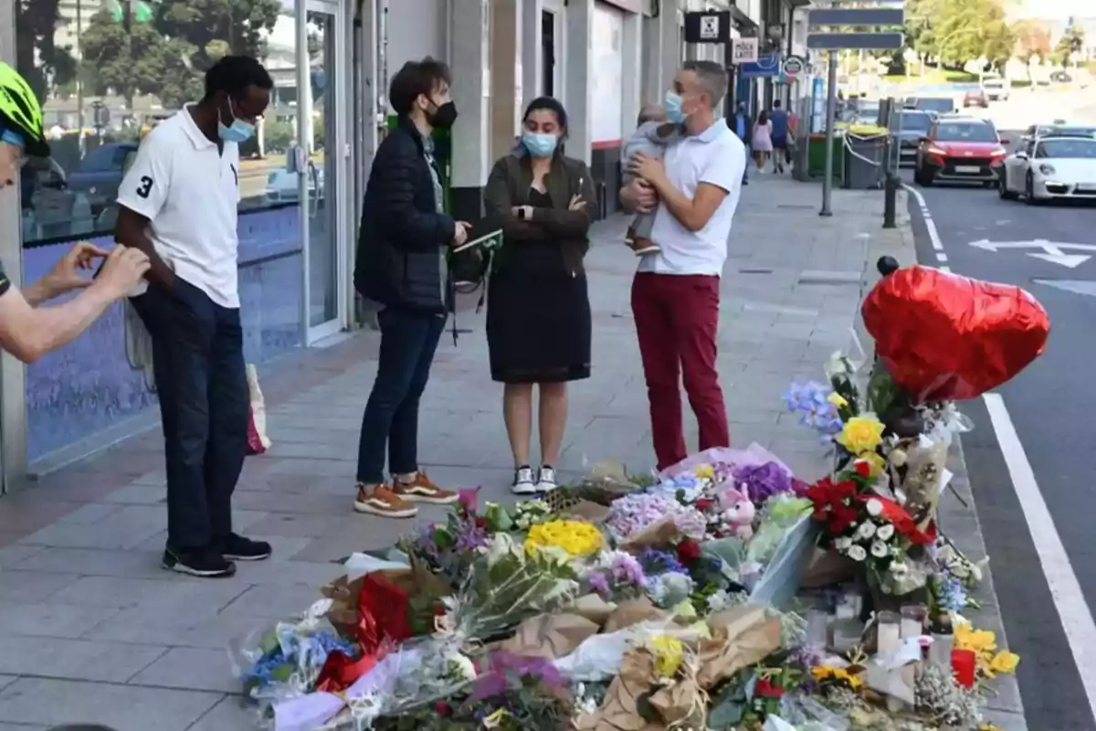 Un grupo de personas con mascarillas se encuentra reunido en una acera junto a un homenaje floral con globos y flores.