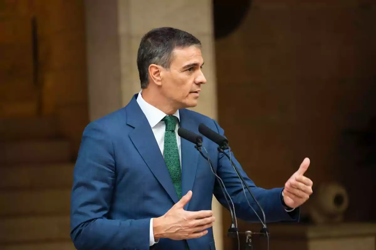 Hombre en traje azul hablando en un micrófono con el pulgar levantado.