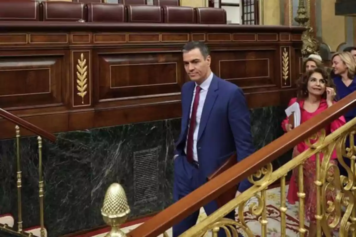 Un hombre con traje azul y corbata roja camina por un pasillo elegante acompañado de varias personas.