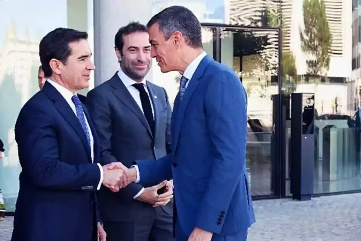 Dos hombres de traje se dan la mano mientras otro hombre observa, todos están de pie frente a un edificio moderno con reflejos en el vidrio.