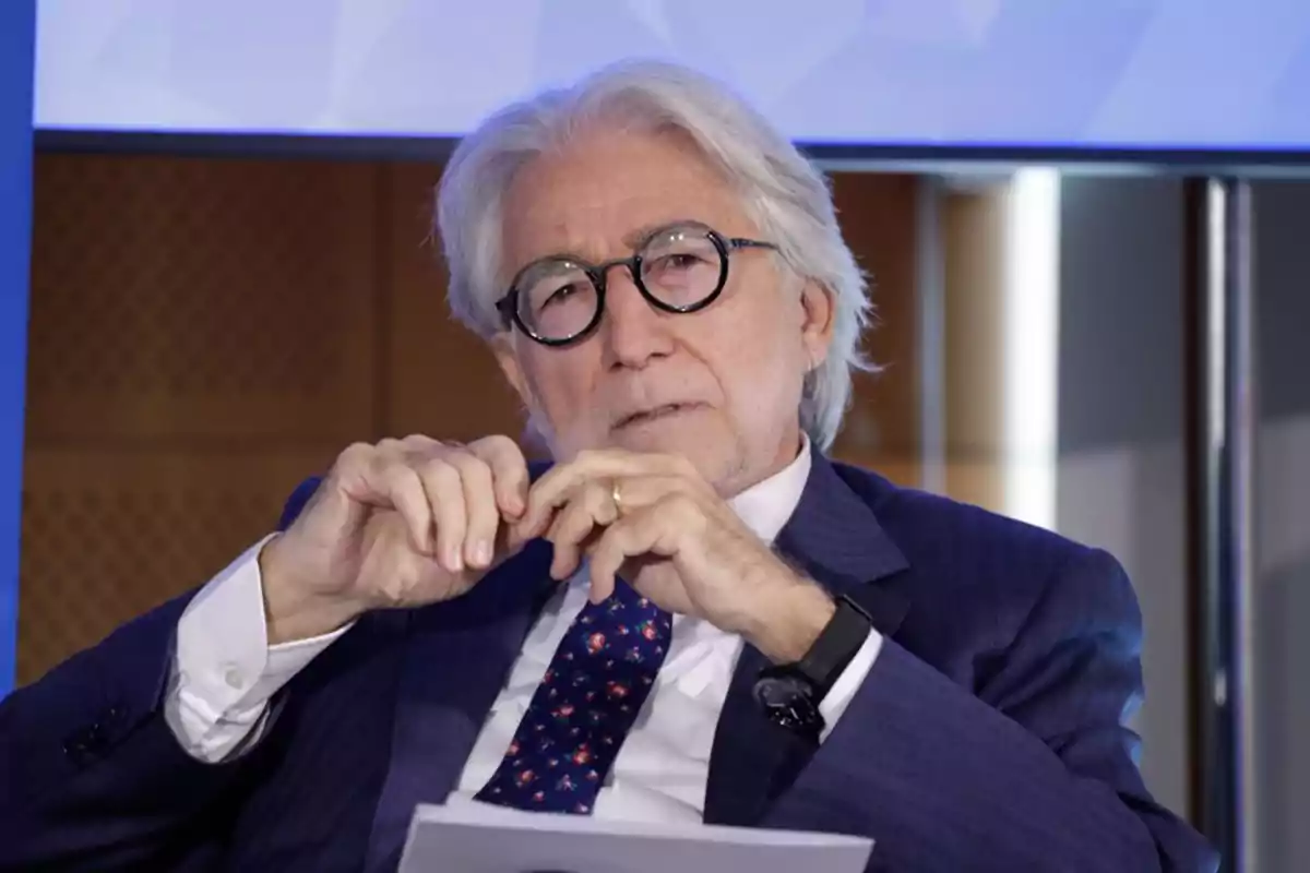 Un hombre de cabello canoso y gafas redondas está sentado, vistiendo un traje azul y una corbata con estampado, mientras sostiene un papel en sus manos. Un hombre de cabello canoso y gafas redondas está sentado, vistiendo un traje azul y una corbata con estampado, mientras sostiene un papel en sus manos.