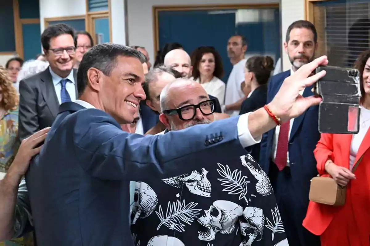 Un hombre con traje y otro con camisa de calaveras se toman una selfie en un evento rodeados de más personas sonrientes Un hombre con traje y otro con camisa de calaveras se toman una selfie en un evento rodeados de más personas sonrientes