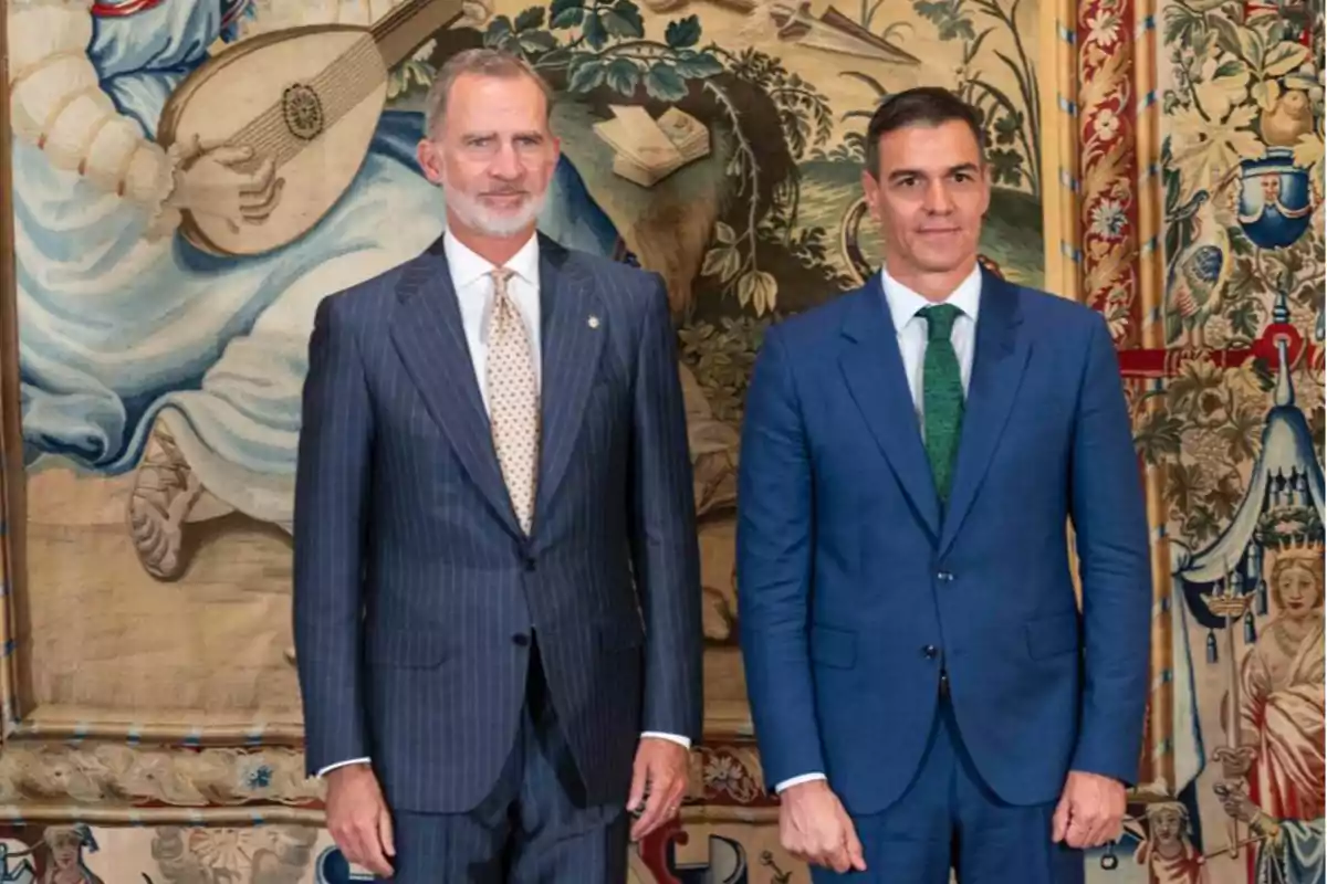 Dos hombres de pie, vestidos con trajes formales, posan frente a un tapiz decorativo.