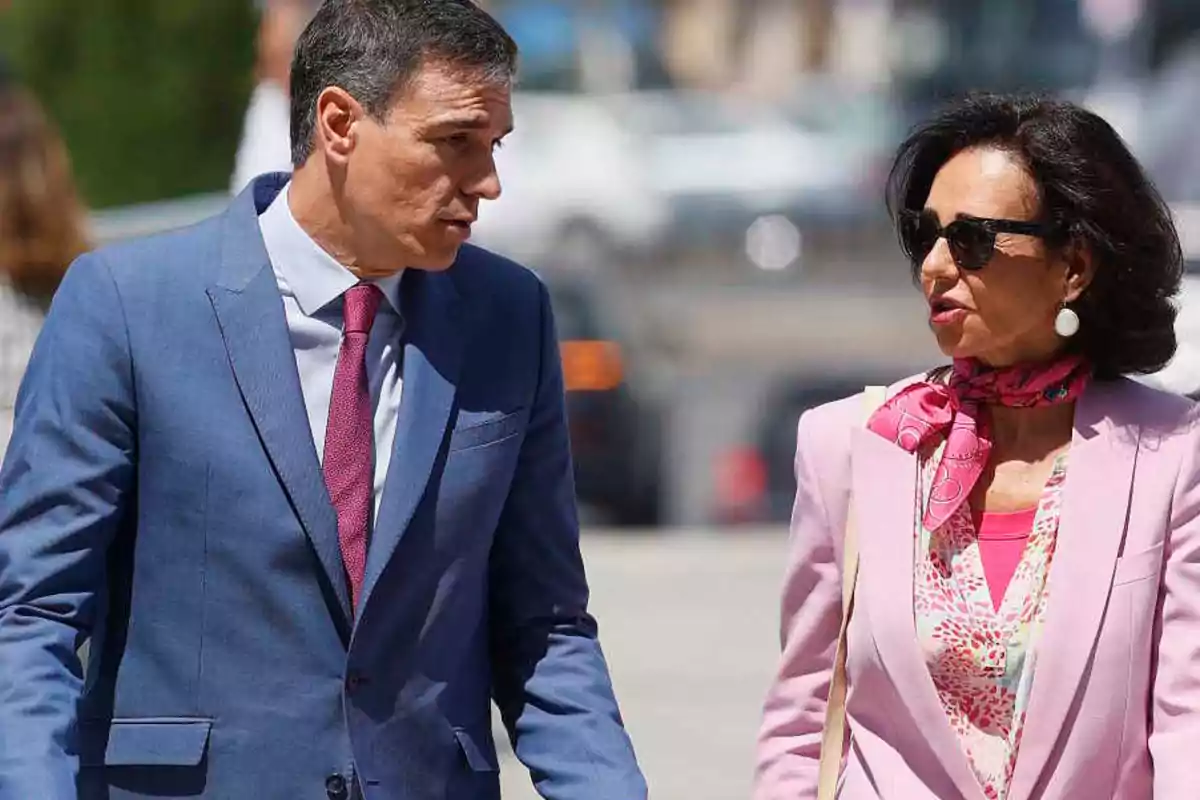 Dos personas conversando mientras caminan, una de ellas lleva un traje azul y corbata roja, y la otra lleva un traje rosa con un pañuelo rosa al cuello y gafas de sol.