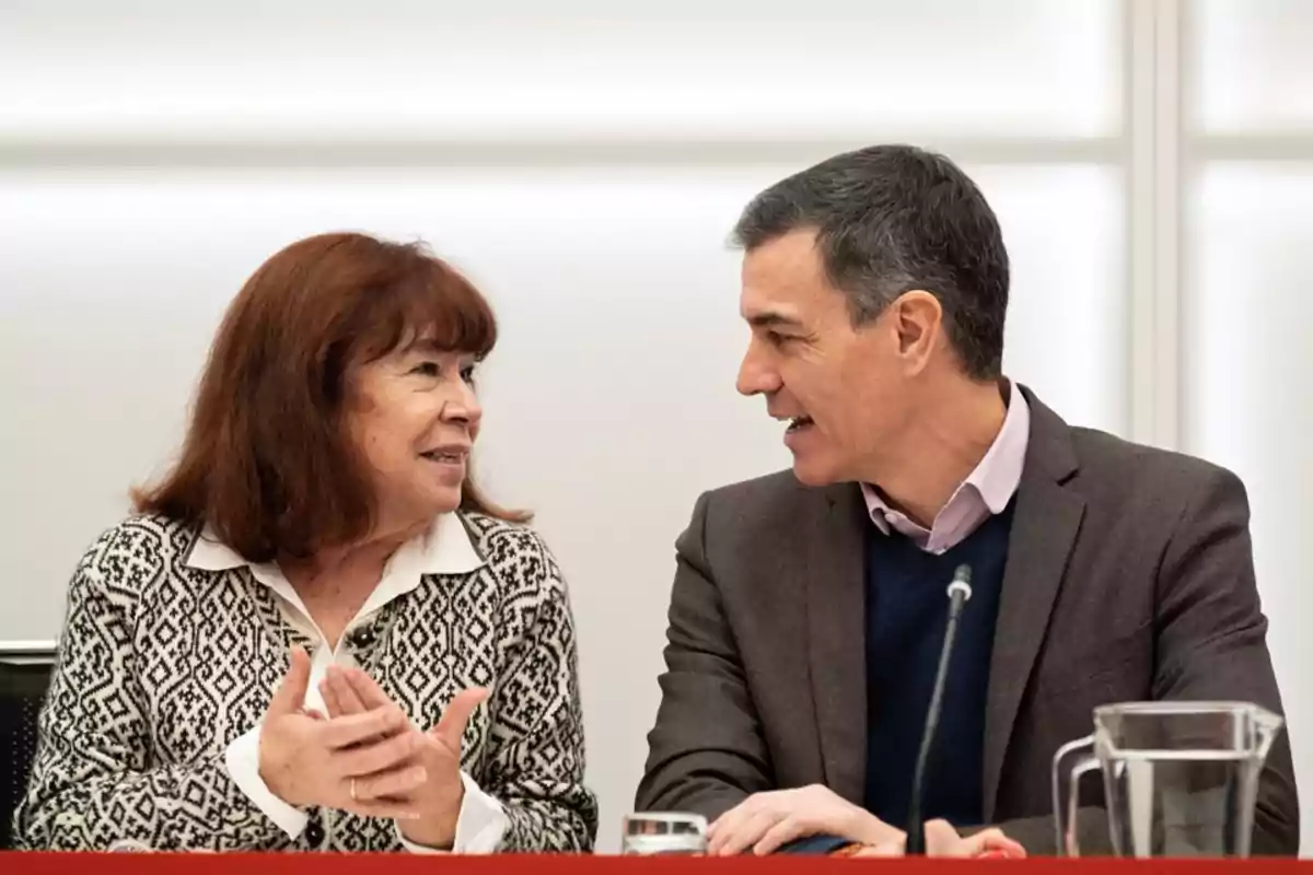 Dos personas sentadas conversando en una mesa durante una reunión, una mujer de cabello castaño y un hombre de cabello corto con chaqueta, ambos sonríen y parecen estar en un ambiente profesional. Dos personas sentadas conversando en una mesa durante una reunión, una mujer de cabello castaño y un hombre de cabello corto con chaqueta, ambos sonríen y parecen estar en un ambiente profesional.