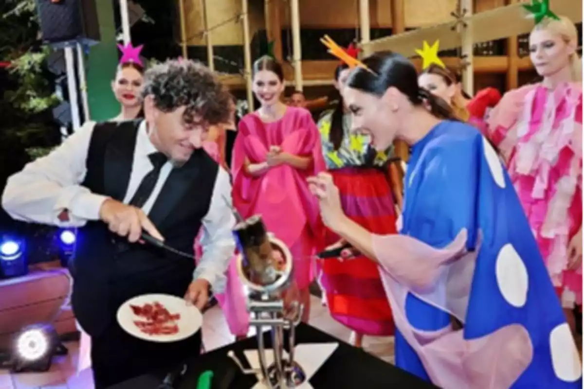 Un hombre cortando jamón mientras una mujer y varias personas con disfraces coloridos observan y sonríen en un evento.