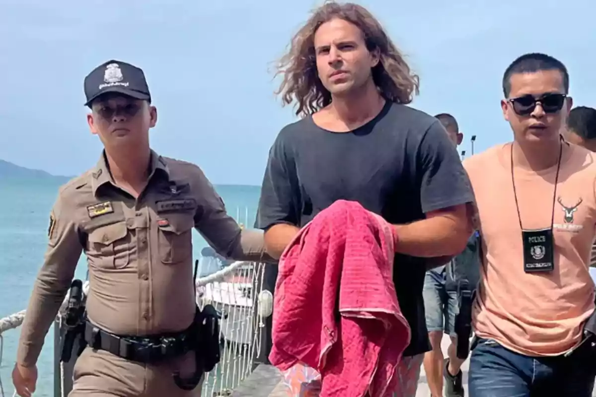 Un hombre escoltado por dos oficiales de policía en un muelle cerca del mar.