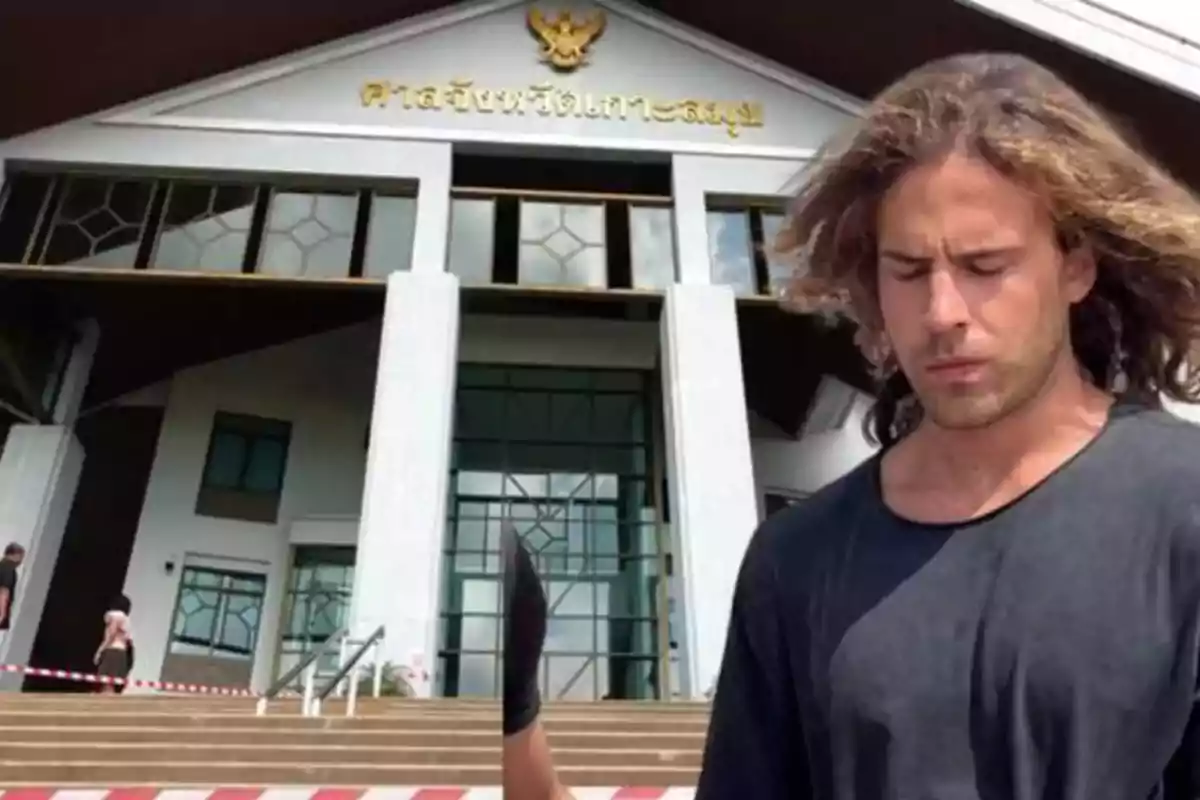 Un hombre de cabello largo y suelto, vestido con una camiseta negra, está de pie frente a un edificio con arquitectura moderna y un letrero en un idioma extranjero.