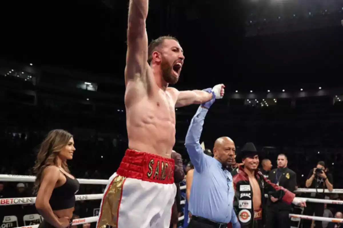 Un boxeador celebra su victoria en el ring con los brazos en alto mientras el árbitro sostiene su mano y el público observa.