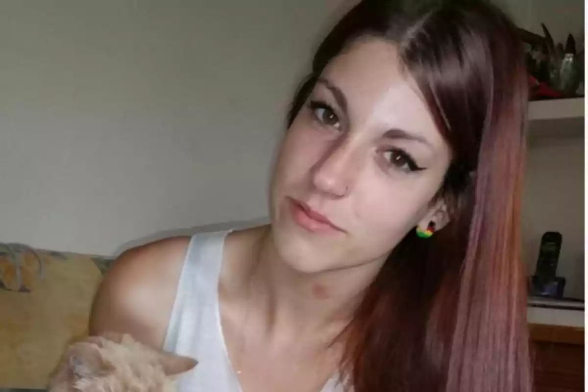 Una mujer con cabello largo y lacio de color castaño claro, usando una camiseta sin mangas de color claro, sosteniendo un gato de color beige.