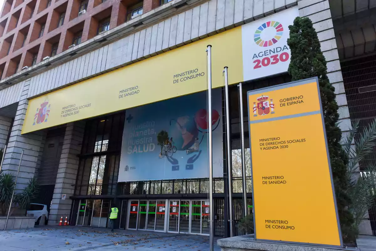 Fachada de un edificio gubernamental en España con carteles del Ministerio de Derechos Sociales y Agenda 2030, Ministerio de Sanidad y Ministerio de Consumo, junto a un letrero de la Agenda 2030 y un cartel que promueve la salud del planeta.