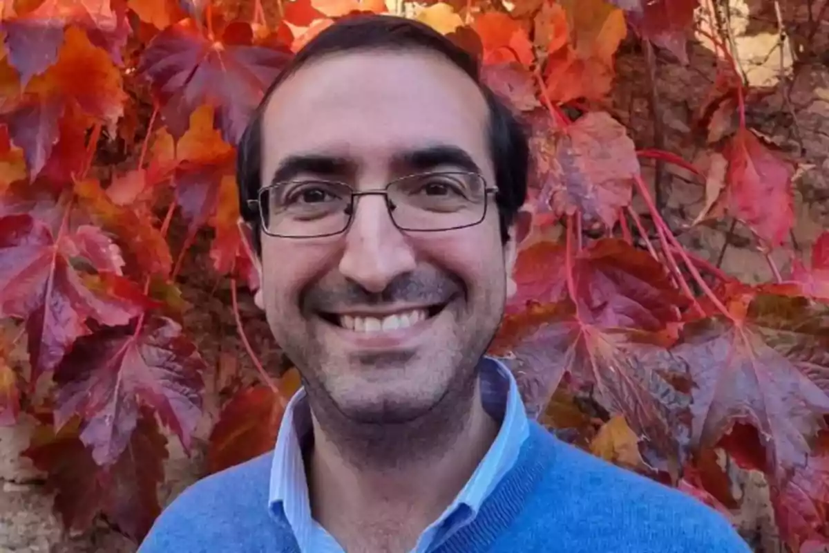 Un hombre sonriente con gafas y suéter azul frente a un fondo de hojas rojas.