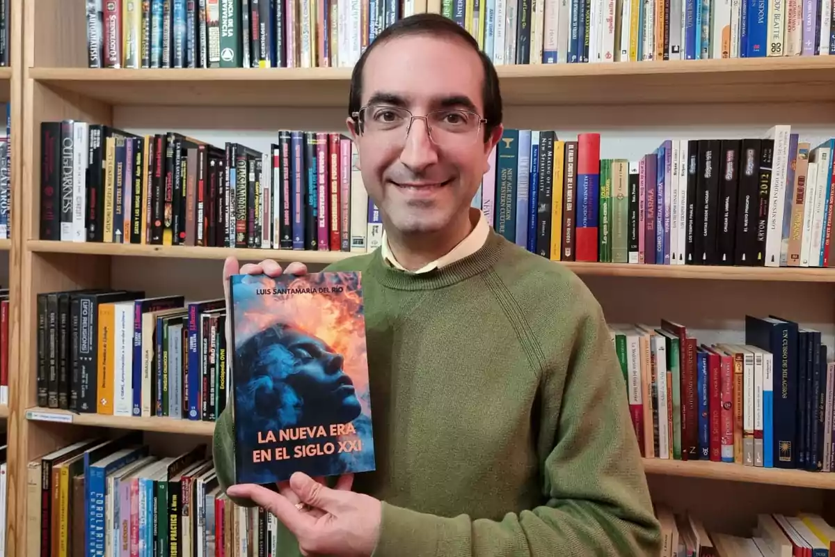 Un hombre con gafas y suéter verde sostiene un libro titulado "La nueva era en el siglo XXI" frente a una estantería llena de libros.