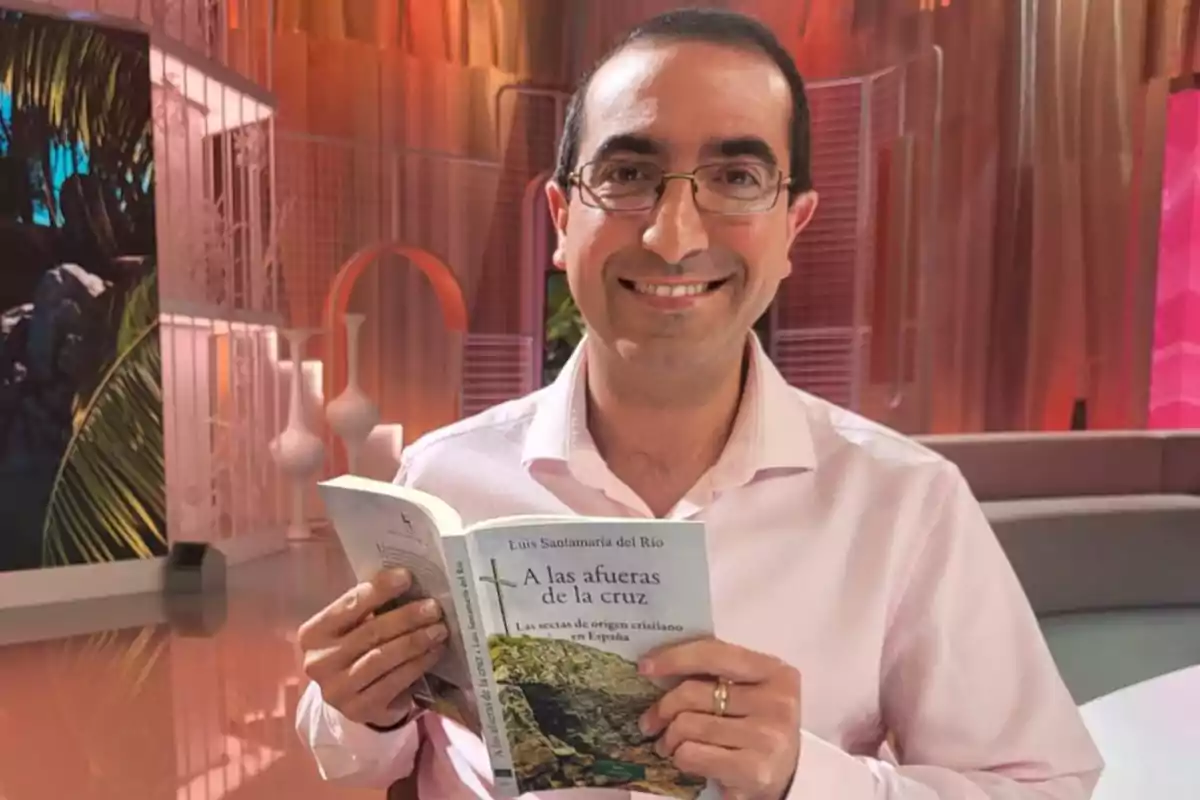 Un hombre con gafas y camisa clara sonríe mientras sostiene un libro titulado 