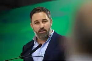 Hombre con barba y cabello corto hablando frente a dos micrófonos con un fondo verde.