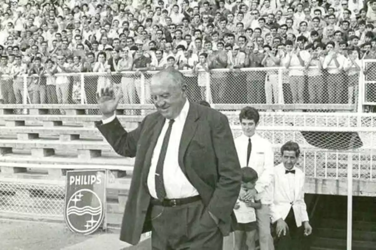 Un hombre mayor con traje oscuro y corbata saluda con la mano mientras camina frente a una multitud que lo aplaude en un estadio deportivo.