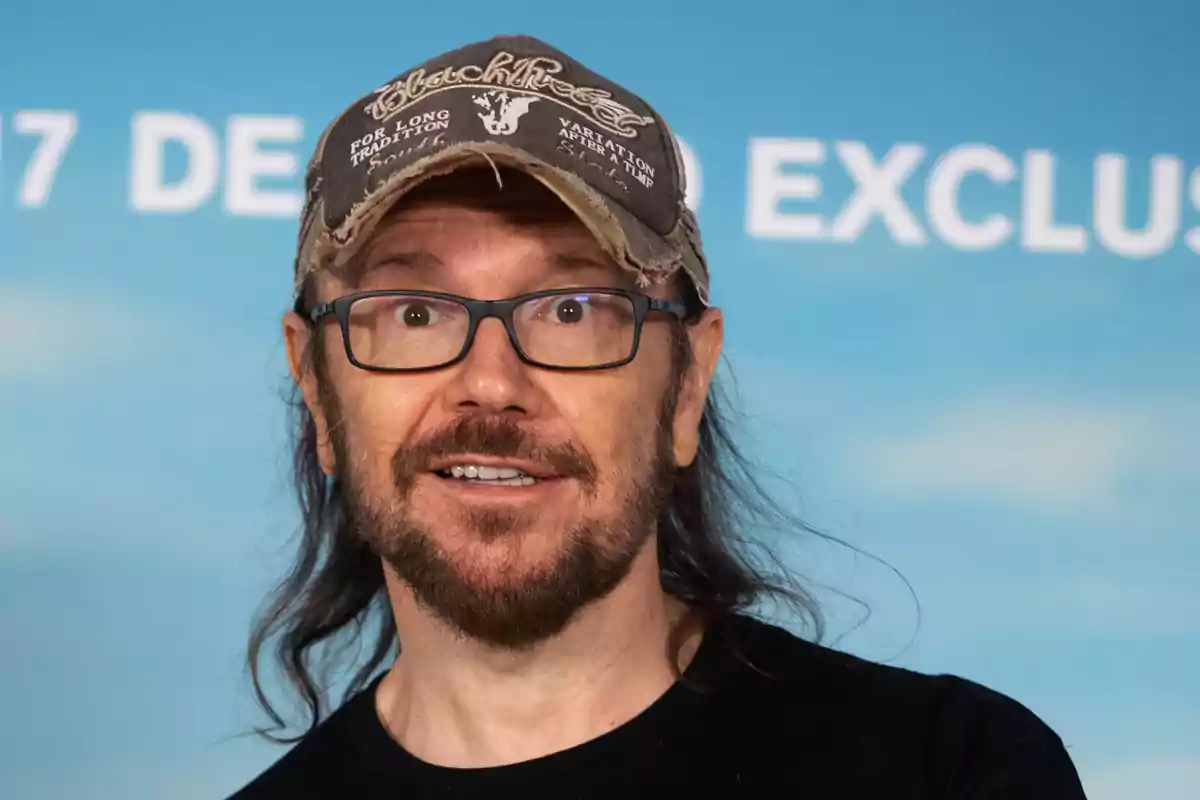 Un hombre con gafas y gorra sonríe frente a un fondo azul con texto parcialmente visible.