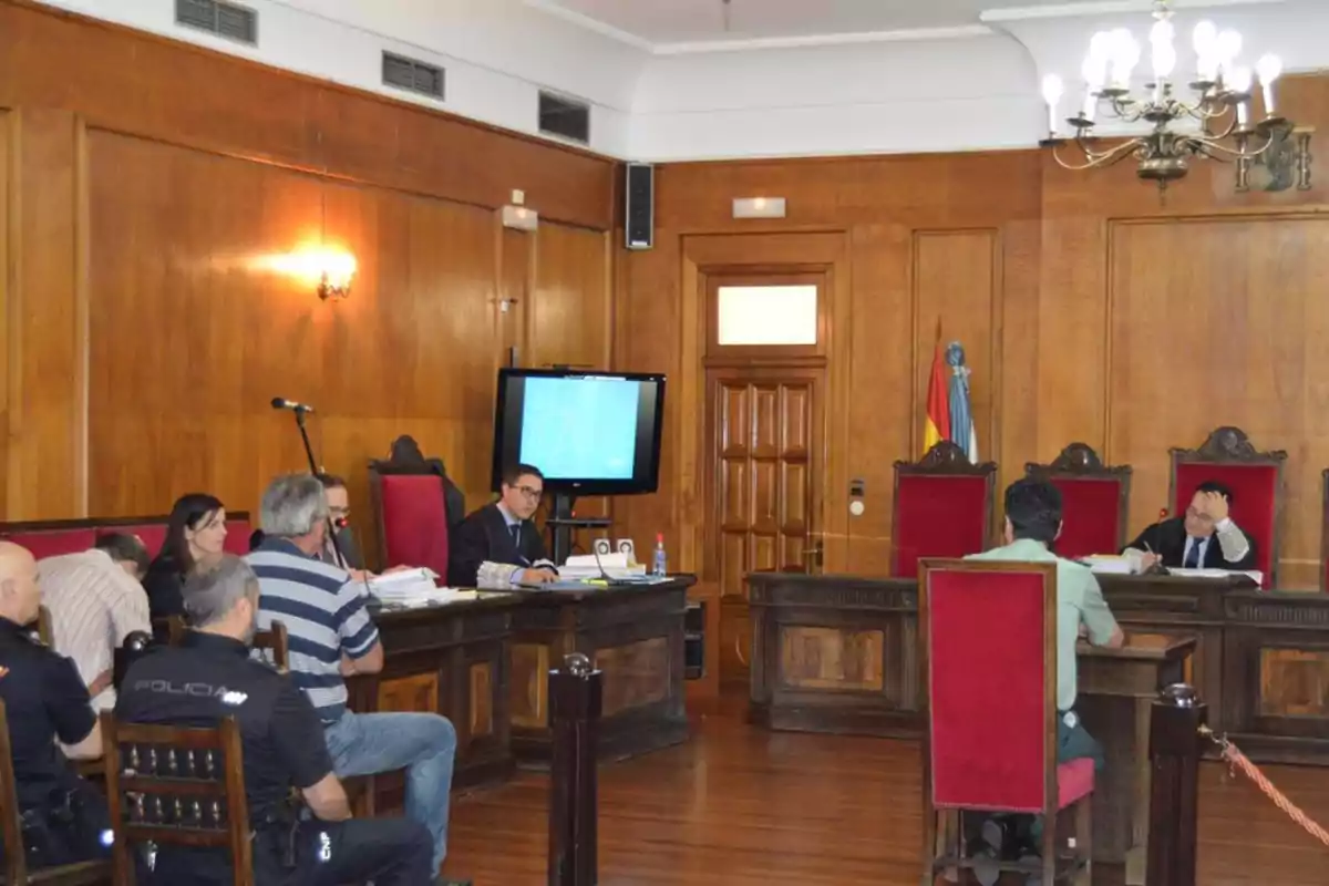 Una sala de tribunal con varias personas presentes, incluyendo jueces y policías, en un ambiente formal con paredes de madera y sillas rojas.