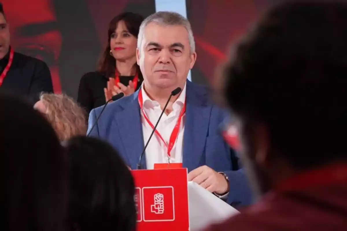 Hombre de cabello canoso con chaqueta azul hablando en un podio rojo frente a un público, con personas aplaudiendo al fondo