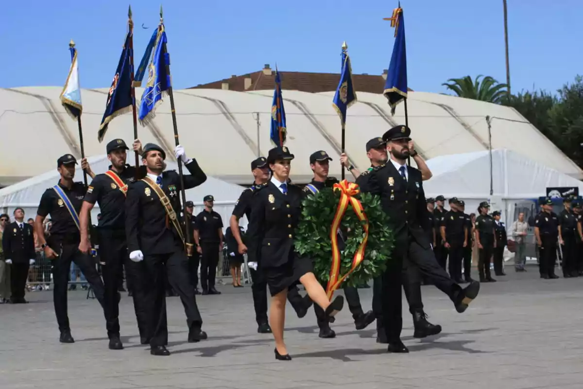 Un grupo de personas uniformadas marcha en formación, llevando banderas y una corona de laurel con una cinta decorativa, en un evento ceremonial al aire libre.