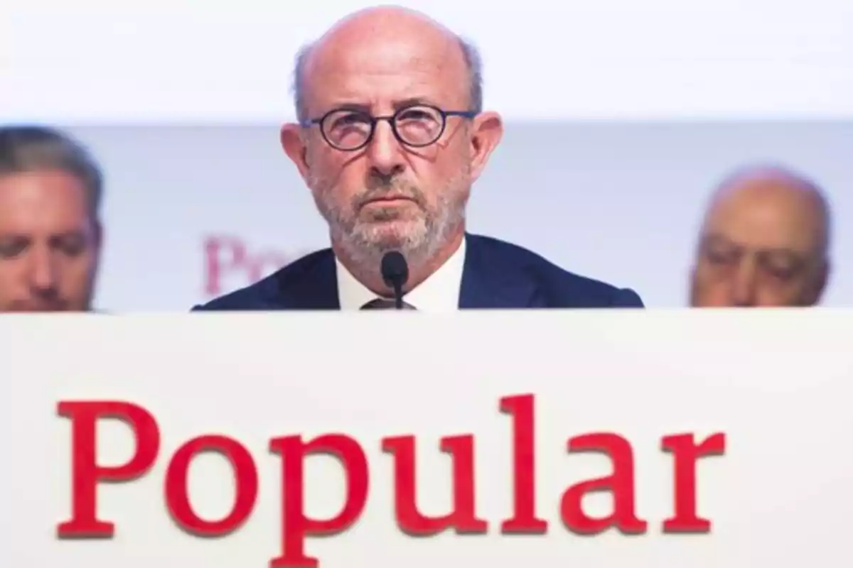 Un hombre con gafas y barba está sentado detrás de un micrófono en un evento con un cartel que dice Un hombre con gafas y barba está sentado detrás de un micrófono en un evento con un cartel que dice