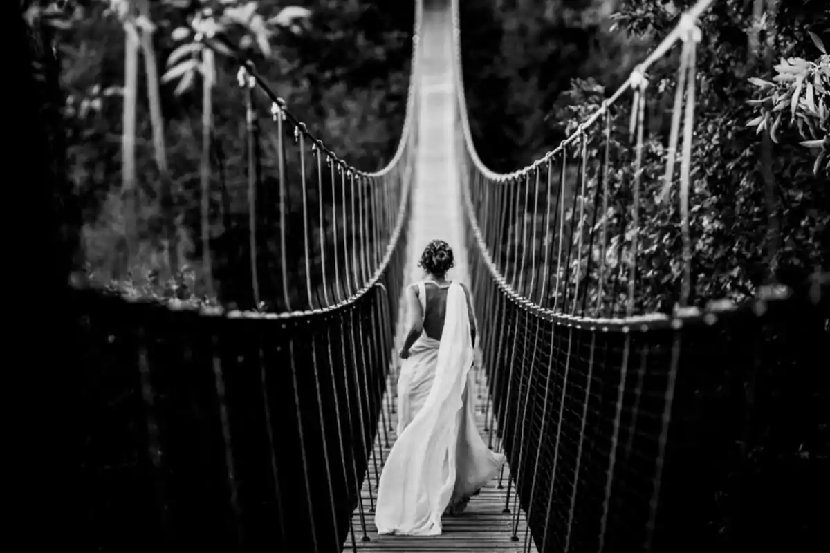 Una persona con un vestido largo y blanco camina por un puente colgante en medio de un entorno natural.