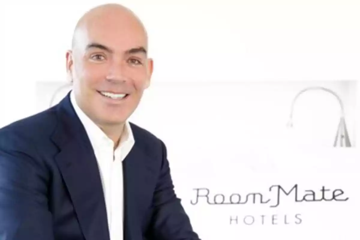 Hombre calvo sonriendo con traje azul oscuro y camisa blanca, con el logo de Room Mate Hotels en el fondo.