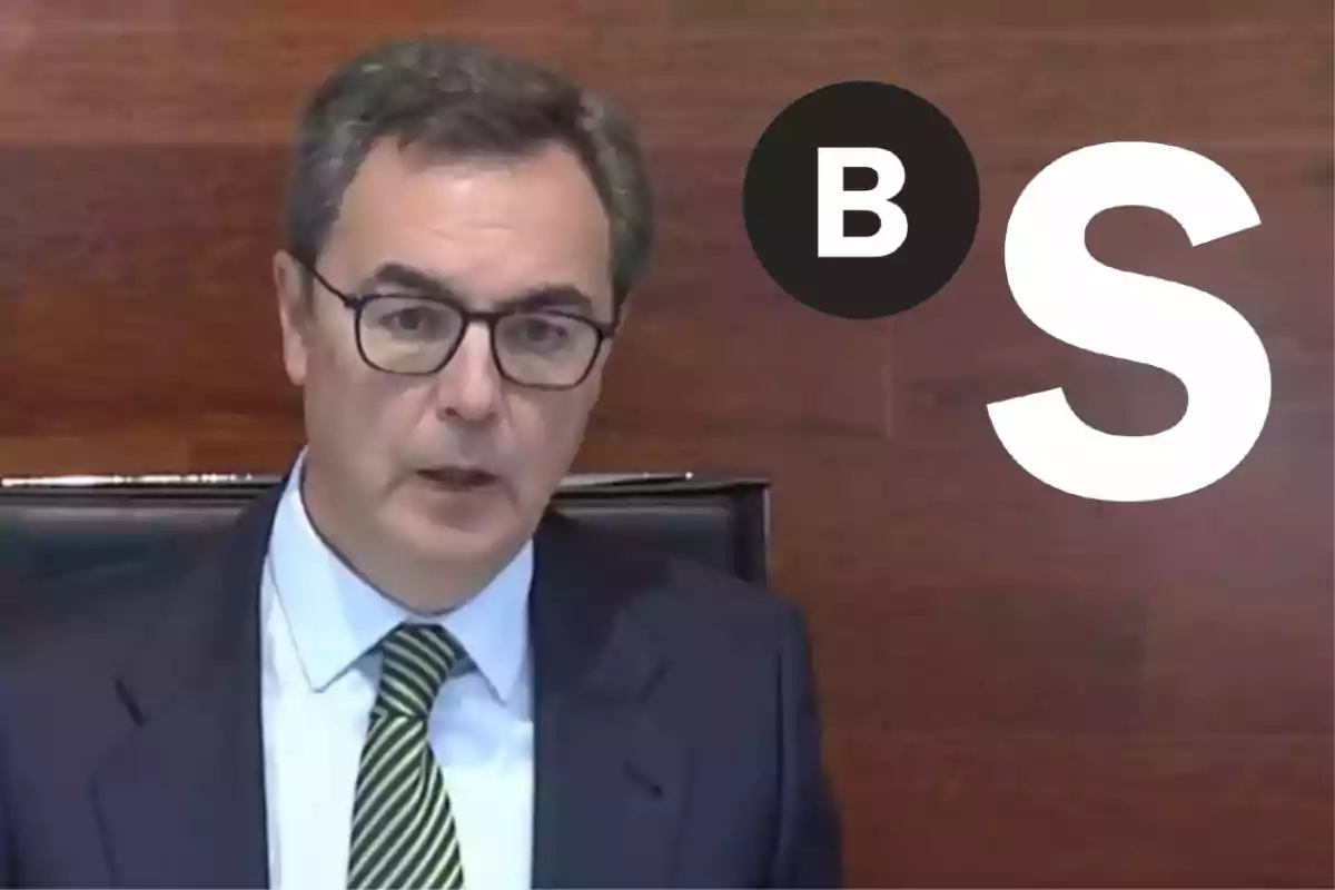Un hombre con gafas y traje oscuro habla frente a un fondo de madera con las letras B y S en el lado derecho.