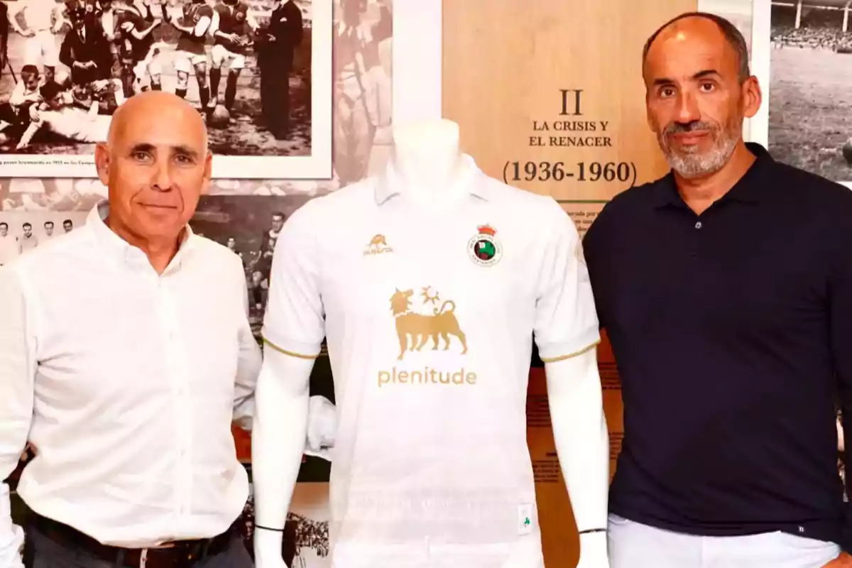 Dos hombres posan junto a un maniquí que lleva una camiseta blanca con logotipos dorados, en un entorno con fotografías históricas en blanco y negro y un cartel que menciona Dos hombres posan junto a un maniquí que lleva una camiseta blanca con logotipos dorados, en un entorno con fotografías históricas en blanco y negro y un cartel que menciona