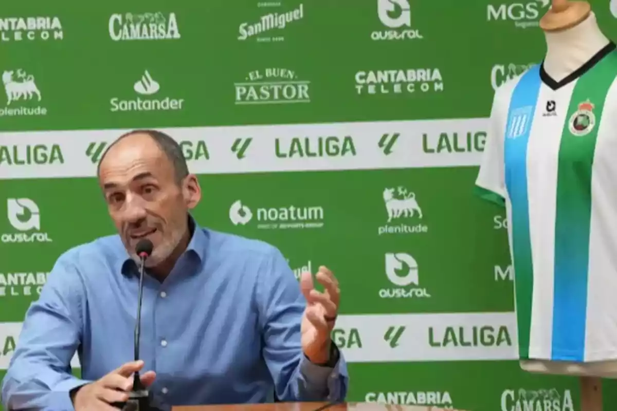 Un hombre hablando en una conferencia de prensa con un micrófono frente a un fondo verde con logotipos de patrocinadores y una camiseta de fútbol en un maniquí a su lado. Un hombre hablando en una conferencia de prensa con un micrófono frente a un fondo verde con logotipos de patrocinadores y una camiseta de fútbol en un maniquí a su lado.