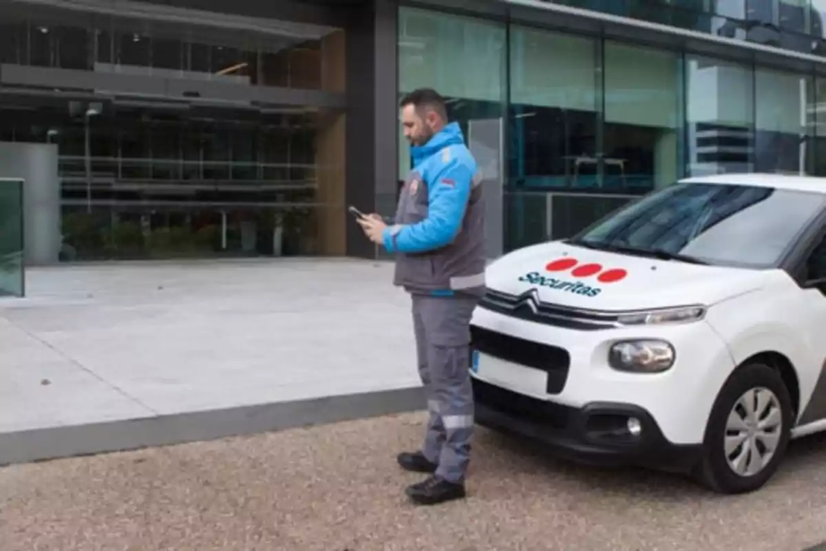 Un guardia de seguridad revisa su dispositivo móvil junto a un vehículo de seguridad frente a un edificio moderno.