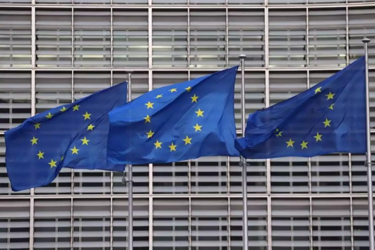 Tres banderas de la Unión Europea ondeando frente a un edificio con ventanas.