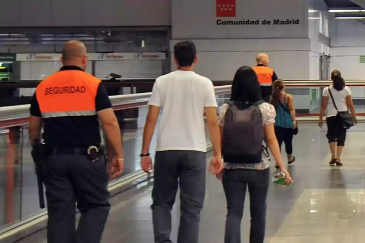 Personas caminando en un pasillo de una estación, acompañadas por personal de seguridad con chalecos naranjas.