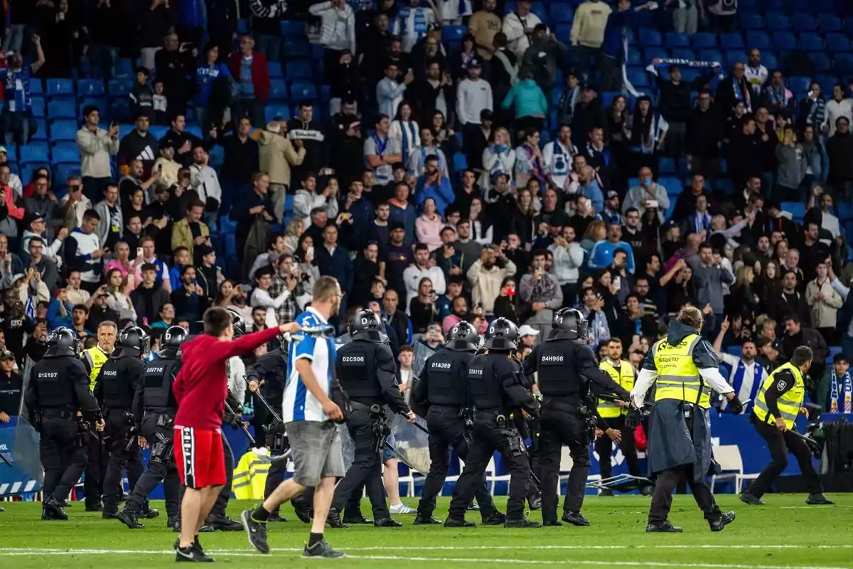 Un grupo de policías y personal de seguridad en un estadio de fútbol mientras la multitud observa desde las gradas.