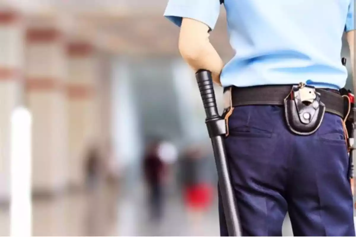 Guardia de seguridad de espaldas con uniforme azul y porra en la mano en un edificio.