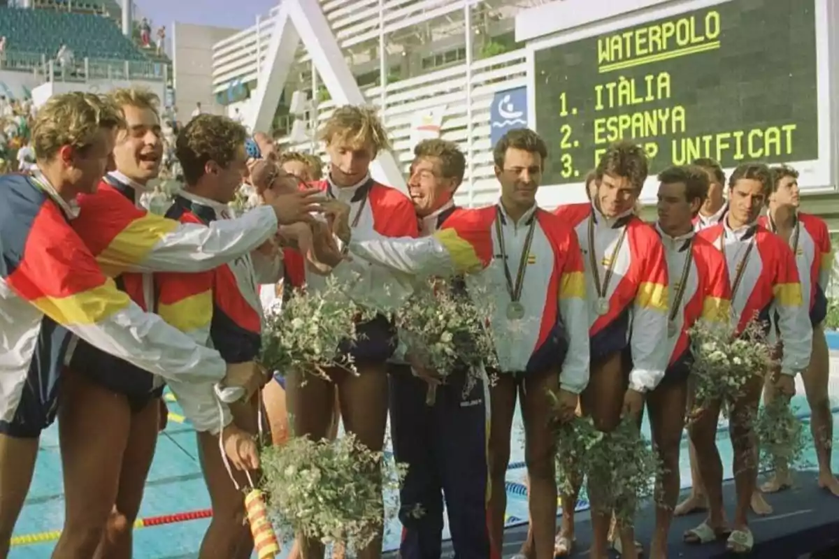 Un equipo de waterpolo celebra con medallas y ramos de flores frente a un marcador que muestra los resultados de una competencia.