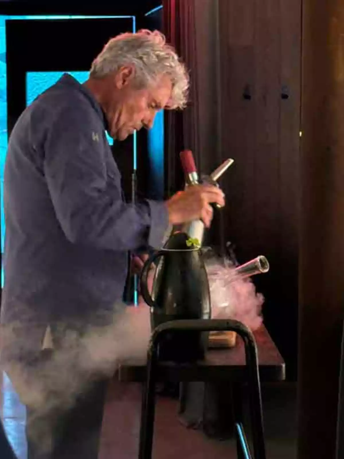 Un hombre de cabello canoso está trabajando con equipo de cocina y una botella, generando vapor en una mesa.