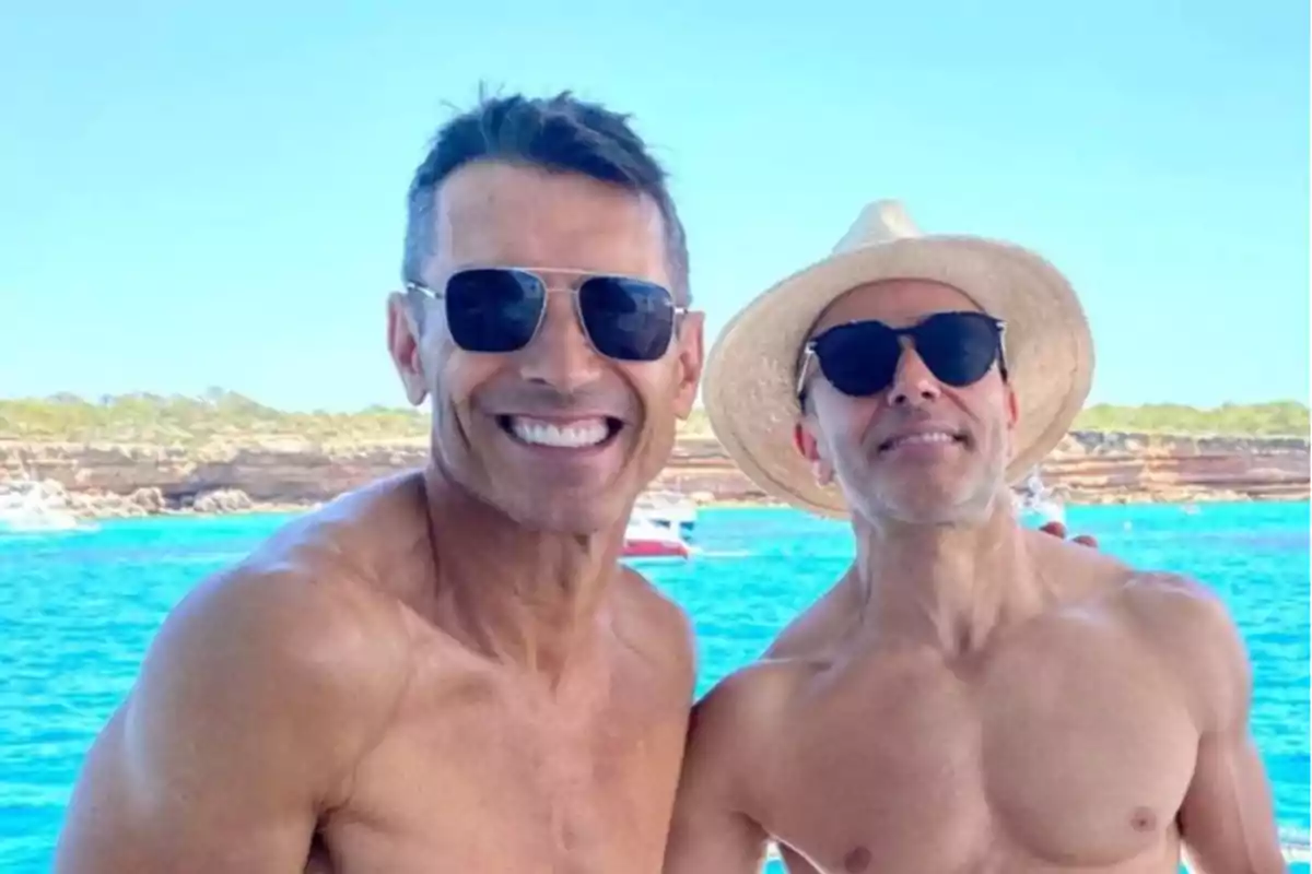 Dos hombres sonrientes posan sin camisa frente al mar con gafas de sol y uno de ellos lleva sombrero de paja Dos hombres sonrientes posan sin camisa frente al mar con gafas de sol y uno de ellos lleva sombrero de paja