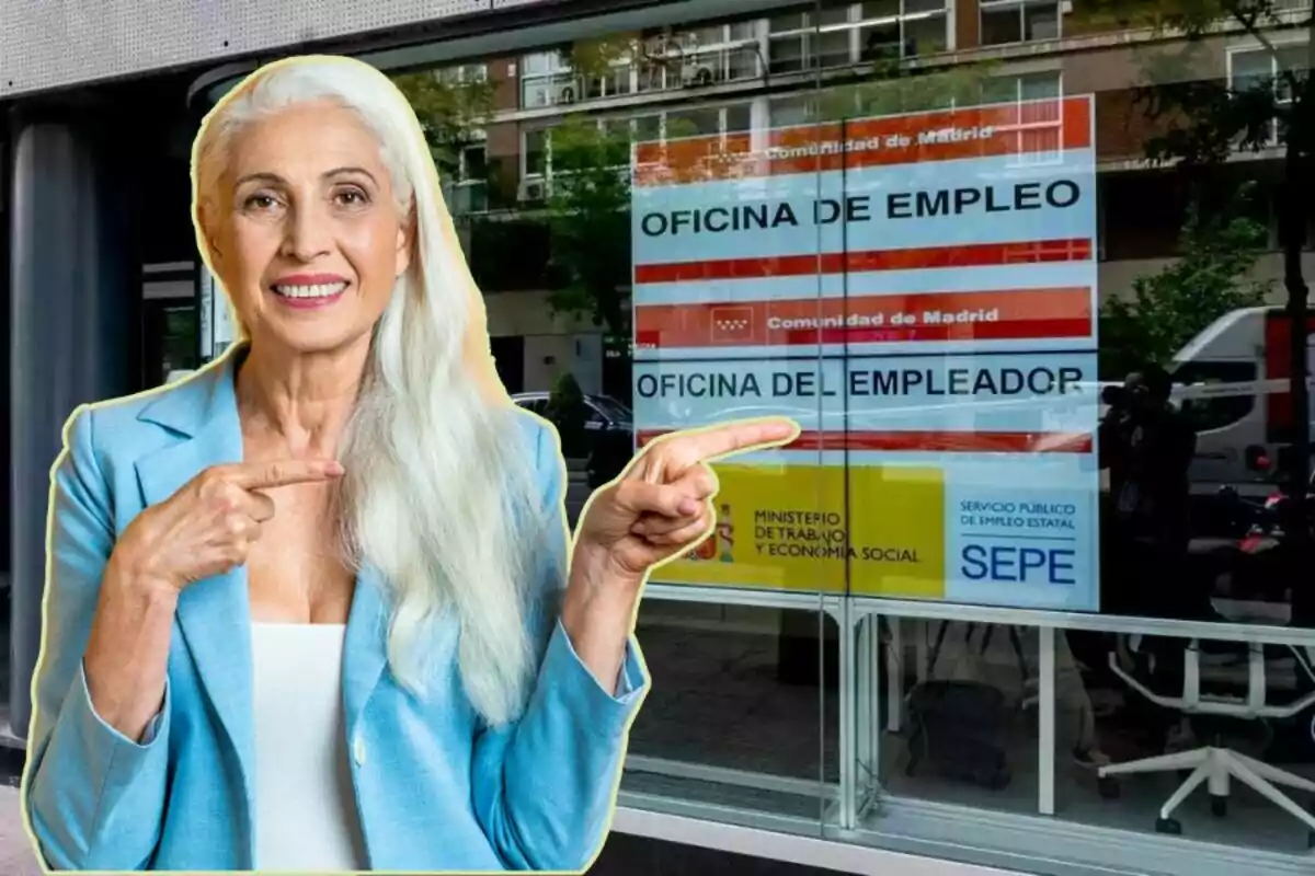 Una mujer de cabello canoso y vestida con un blazer azul claro está sonriendo y señalando con ambos dedos índices hacia un cartel de la Oficina de Empleo de la Comunidad de Madrid, visible a través de una ventana.