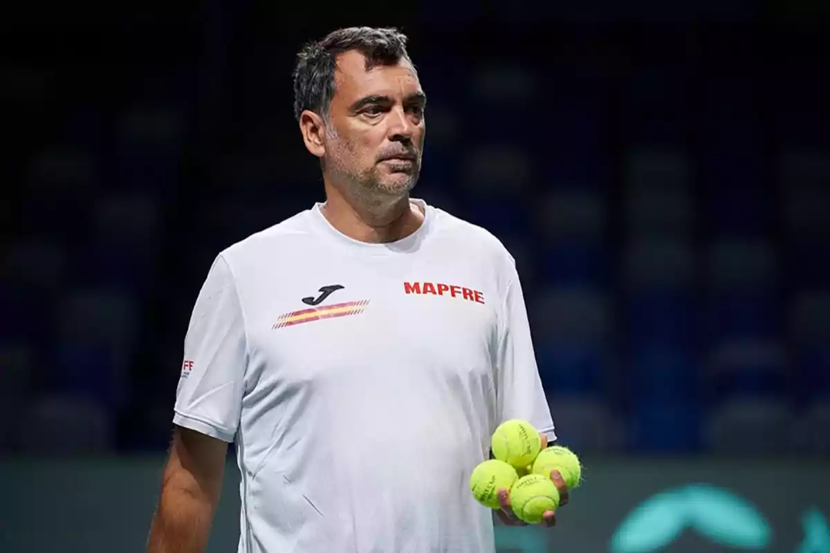 Un hombre con camiseta blanca sostiene varias pelotas de tenis en una cancha.