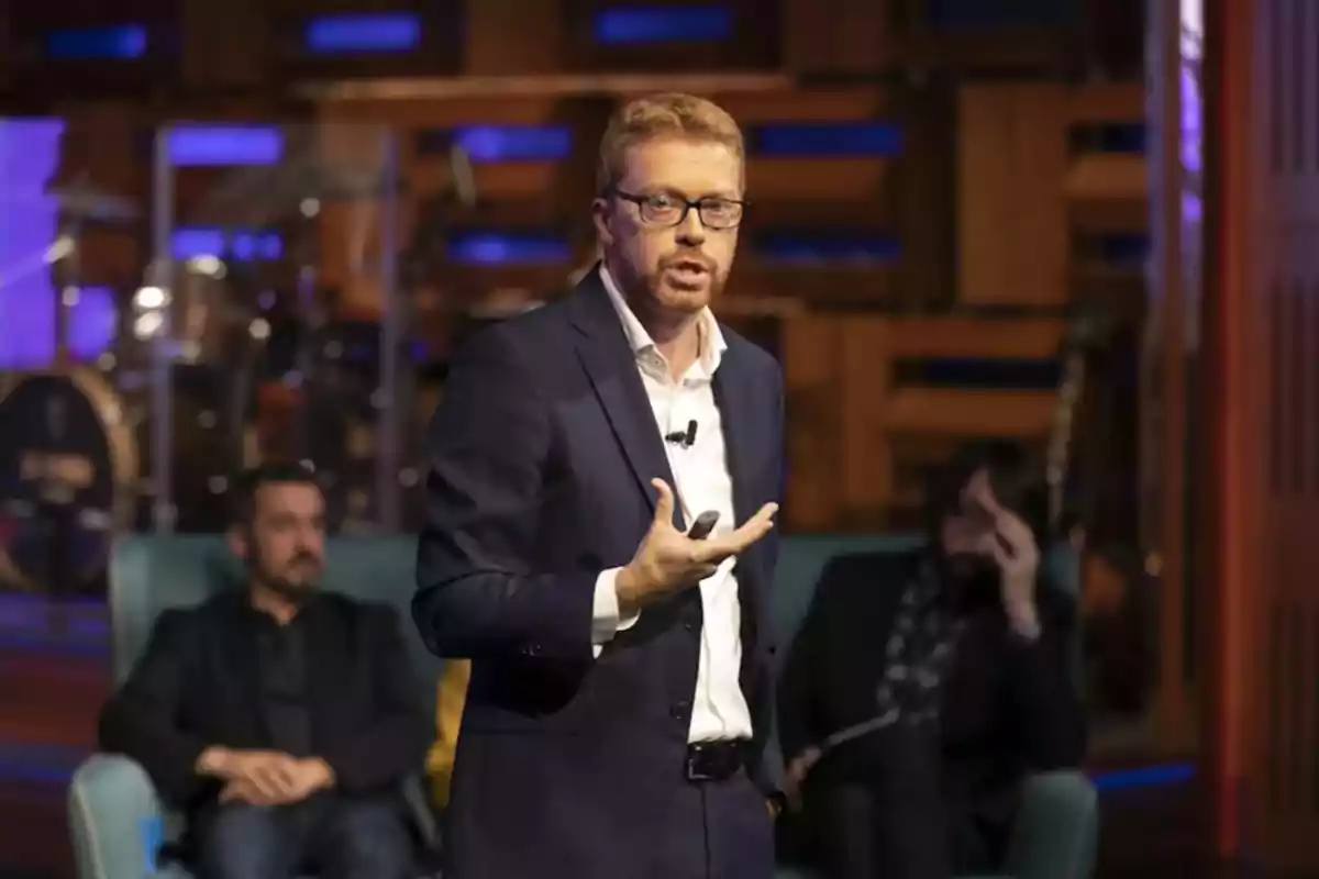 Un hombre con gafas y traje oscuro está hablando en un escenario, mientras dos personas están sentadas en el fondo.