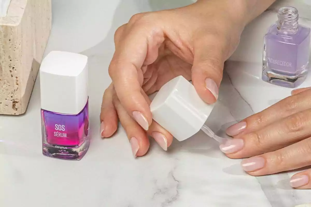 Mano aplicando esmalte transparente en las uñas sobre una superficie de mármol junto a frascos de productos para el cuidado de uñas Mano aplicando esmalte transparente en las uñas sobre una superficie de mármol junto a frascos de productos para el cuidado de uñas