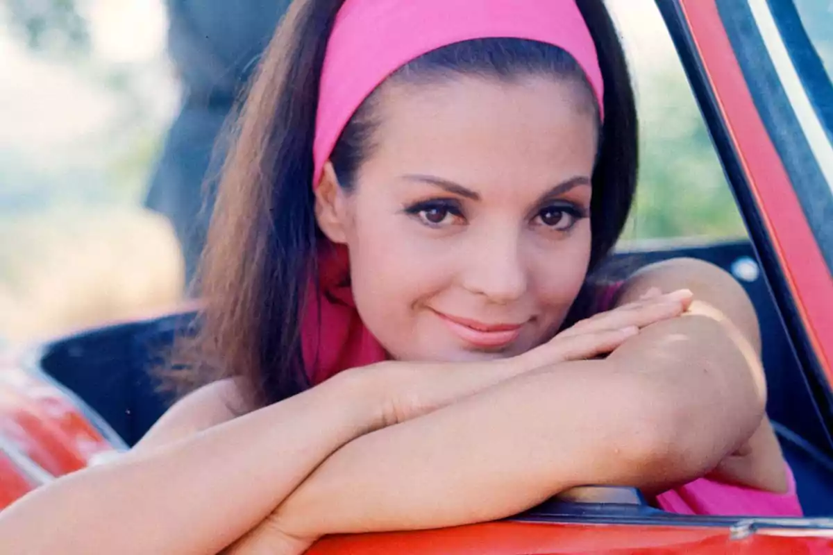 Mujer con cinta rosa en el cabello sonriendo y apoyada en un auto rojo.