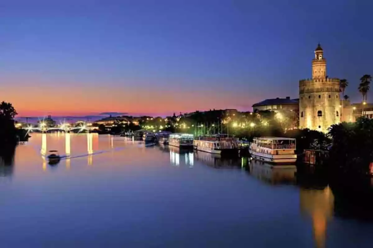 Vista nocturna de un río con barcos y una torre iluminada al fondo bajo un cielo al atardecer.