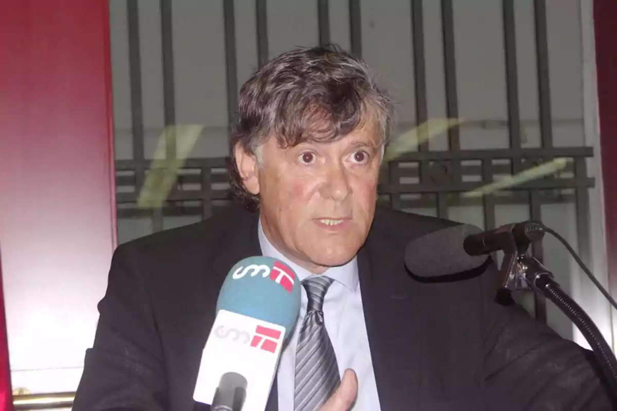 Hombre de cabello canoso y traje oscuro hablando frente a varios micrófonos en una conferencia de prensa Hombre de cabello canoso y traje oscuro hablando frente a varios micrófonos en una conferencia de prensa