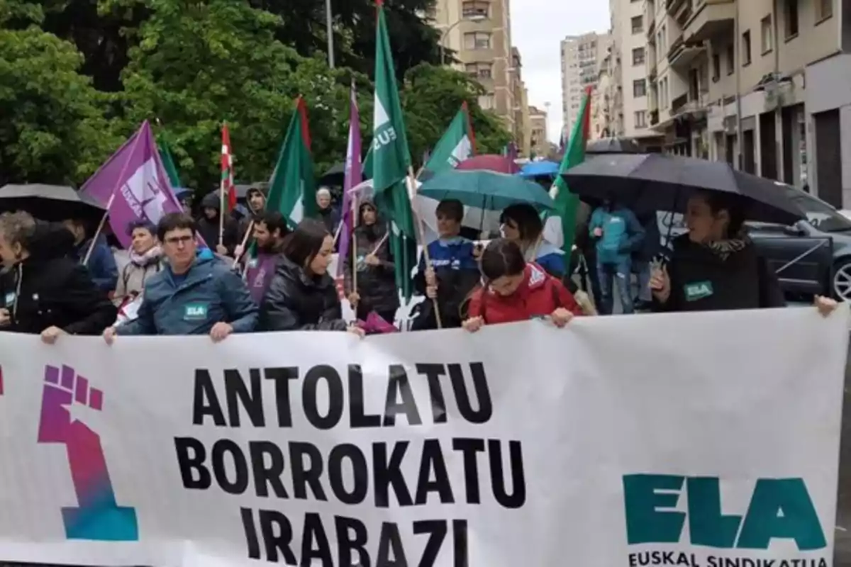 Personas marchando en una manifestación con pancartas y banderas bajo la lluvia en una calle urbana.