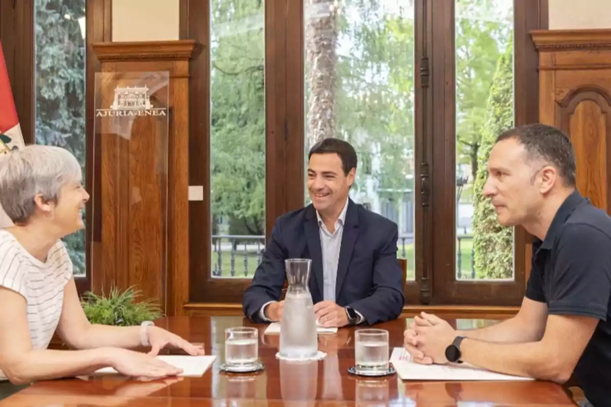 Tres personas sentadas alrededor de una mesa de madera en una reunión, con ventanas grandes y un entorno verde visible al fondo.