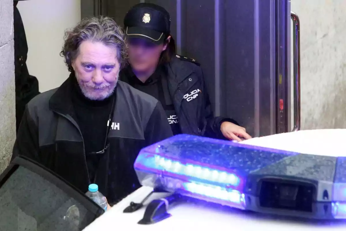 Un hombre con cabello canoso y barba es escoltado por una oficial de policía mientras sale de un edificio, con una luz azul de una patrulla en primer plano.