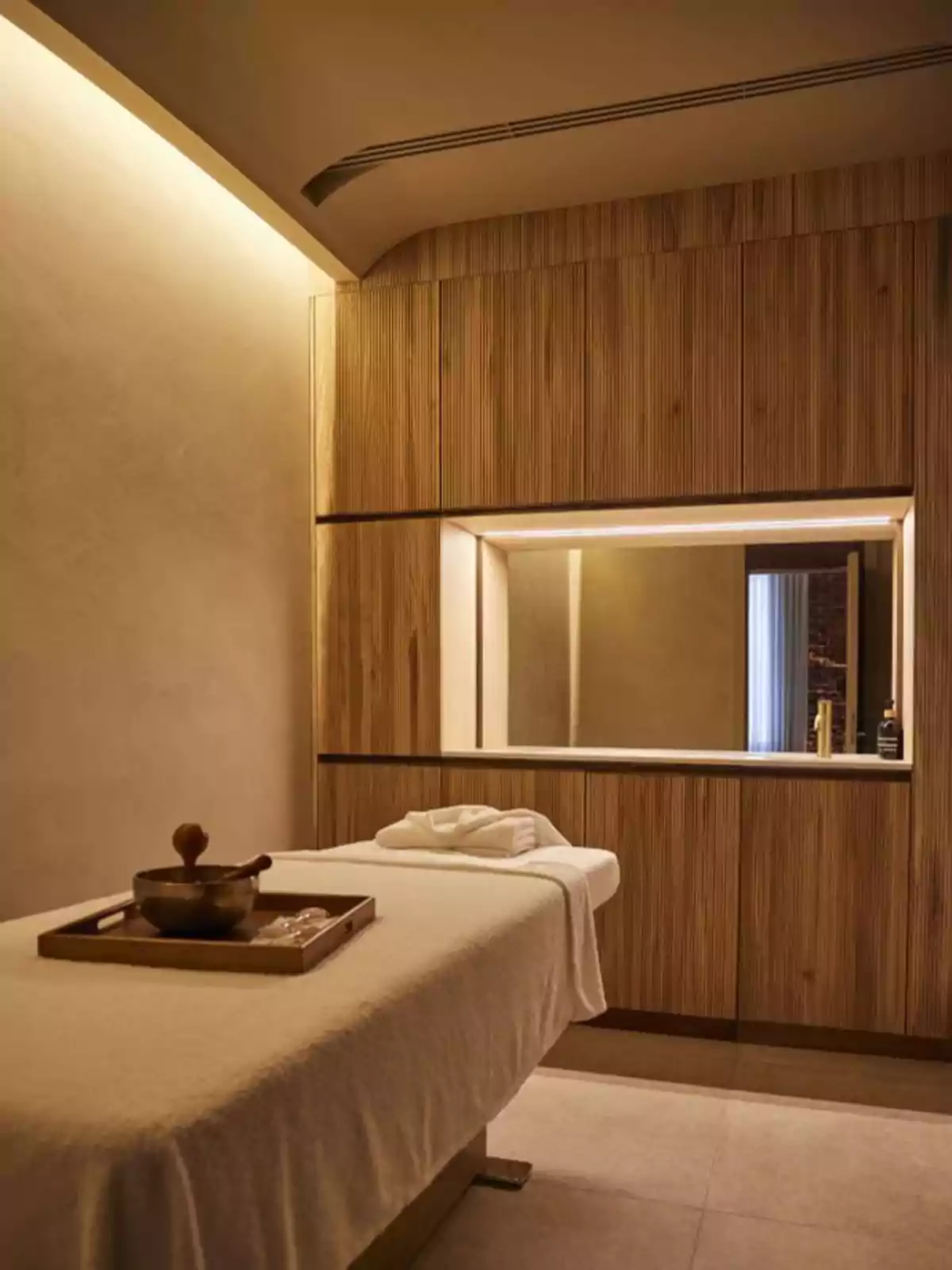 Sala de masajes con iluminación cálida, mesa de masaje cubierta con una toalla blanca y una bandeja con utensilios sobre ella, paredes y muebles de madera.